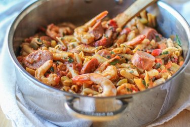 Stainless steel pot full of "gumbo" pasta on a stove top, stirred by a wooden spoon.