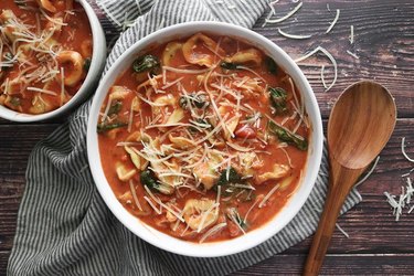 Overhead view of tortellini soup in white bowls, with a wooden spoon and kitchen towel alongside.